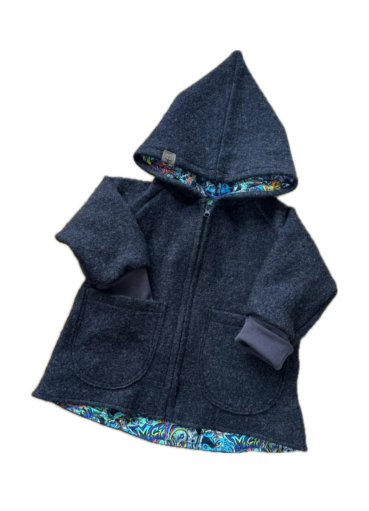 Eine dunkelgraue Jacke, die liegend fotografiert wurde. Die Kinder Jacke ist aus Wolle mit Reißverschluss. Die Jacke hat eine Zipfelmütze und es in grau mit Graffiti Design Futter innen vernäht.